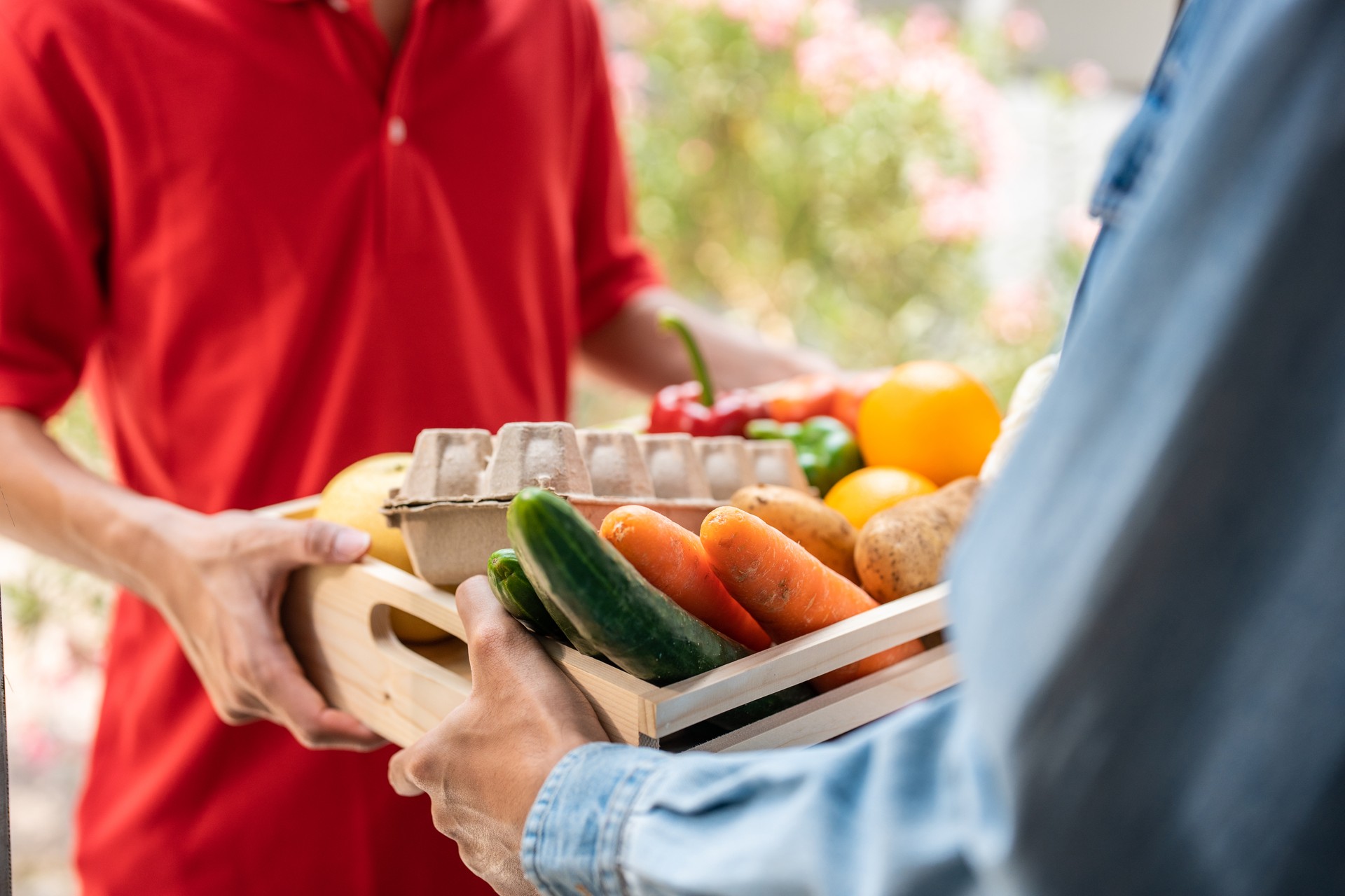 Close up hands of young delivery man delivering package to customer. Attractive postman in red uniform working, deliver a box of fruit and vegetable groceries to buyer infront of the door at home.